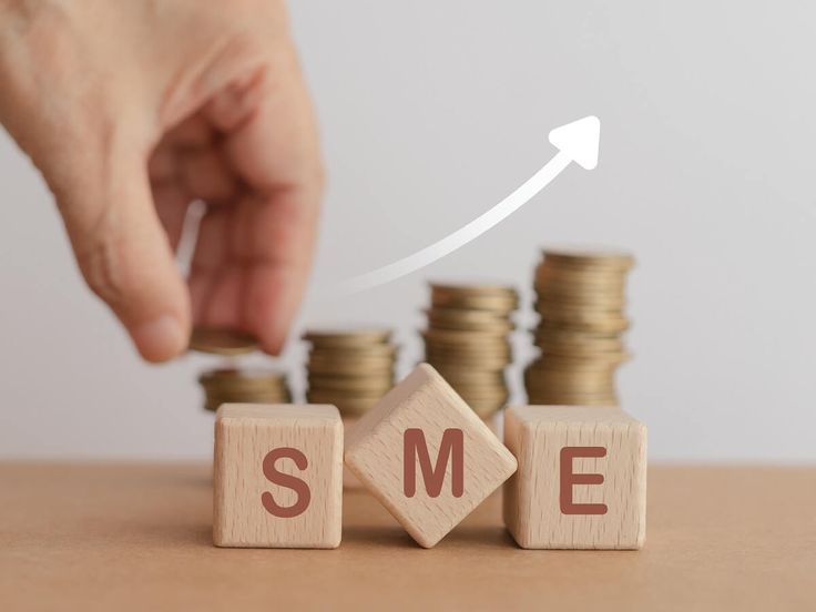A small business growing, represented by wooden SME blocks with a hand stacking coins beside a rising arrow
